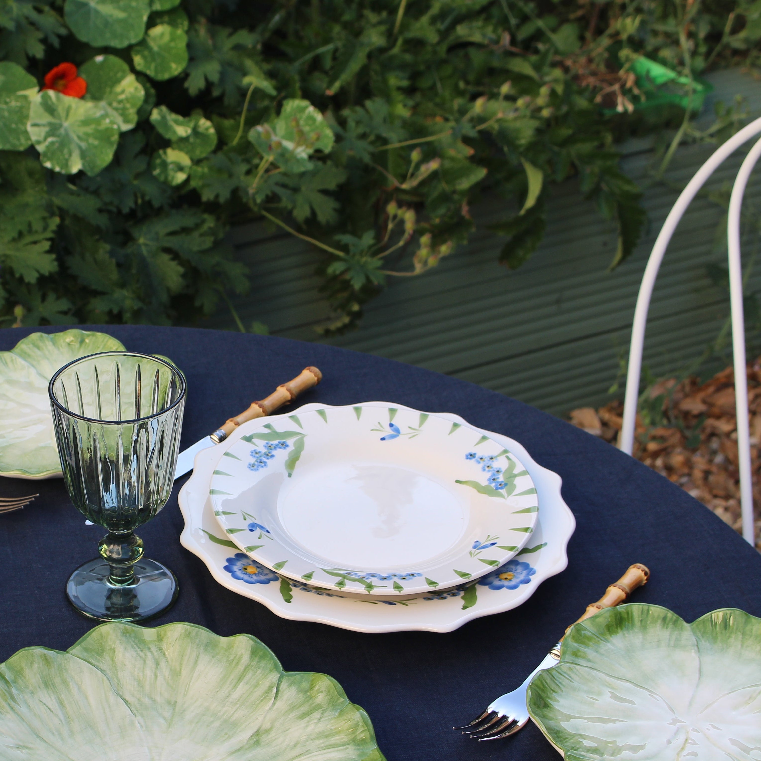 A styled outdoor table setting with leaf-shaped plates, blue and green glass goblets, and gold-handled cutlery on a navy tablecloth, with garden foliage in the background.