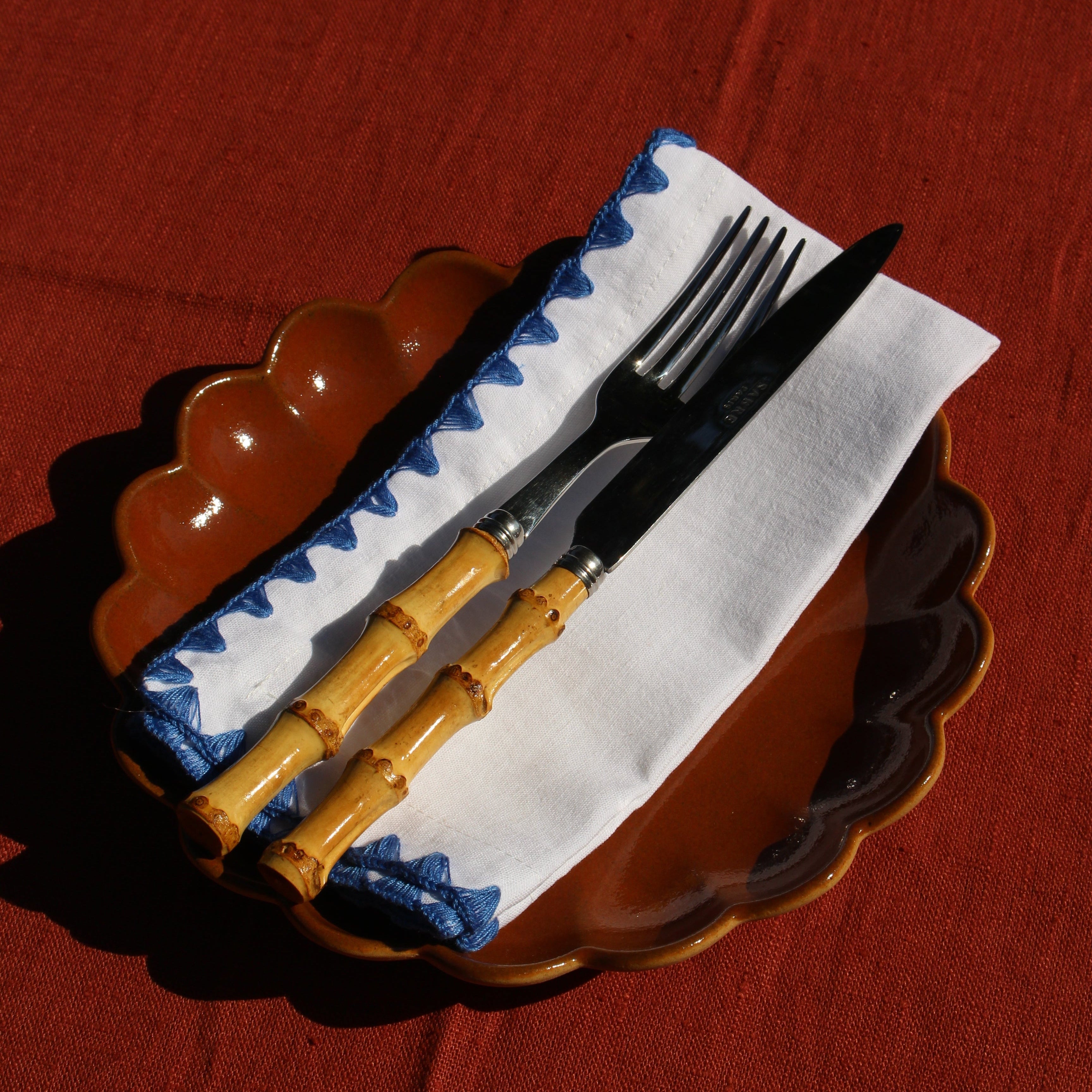 A set of cutlery with light wood bamboo-style handles and black blades, resting on a white napkin with blue zigzag trim, placed on a scalloped brown plate over a red tablecloth.