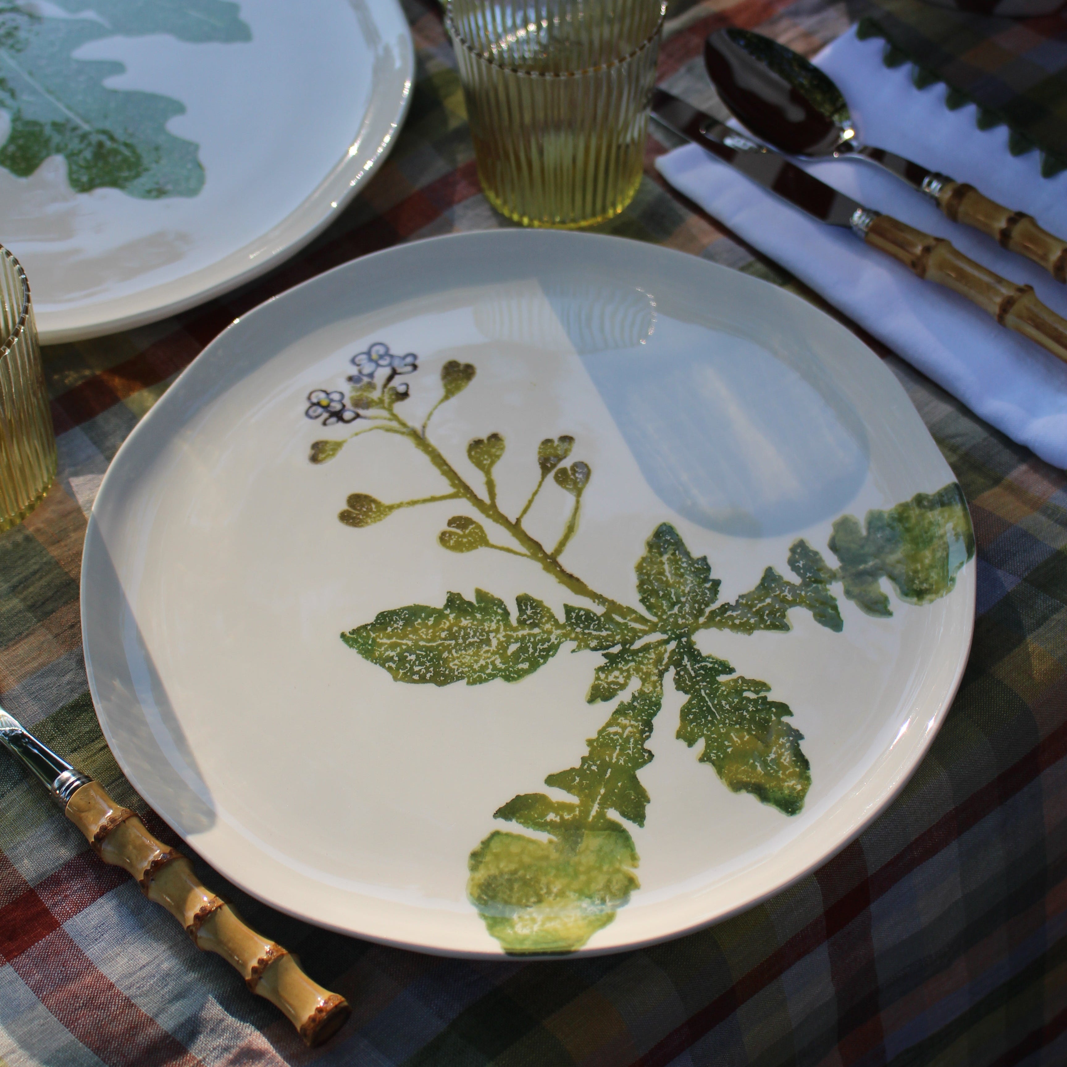 A white ceramic plate with a green leafy botanical print, styled on a rustic outdoor table with bamboo-handled cutlery, a striped plaid tablecloth, and other matching tableware.