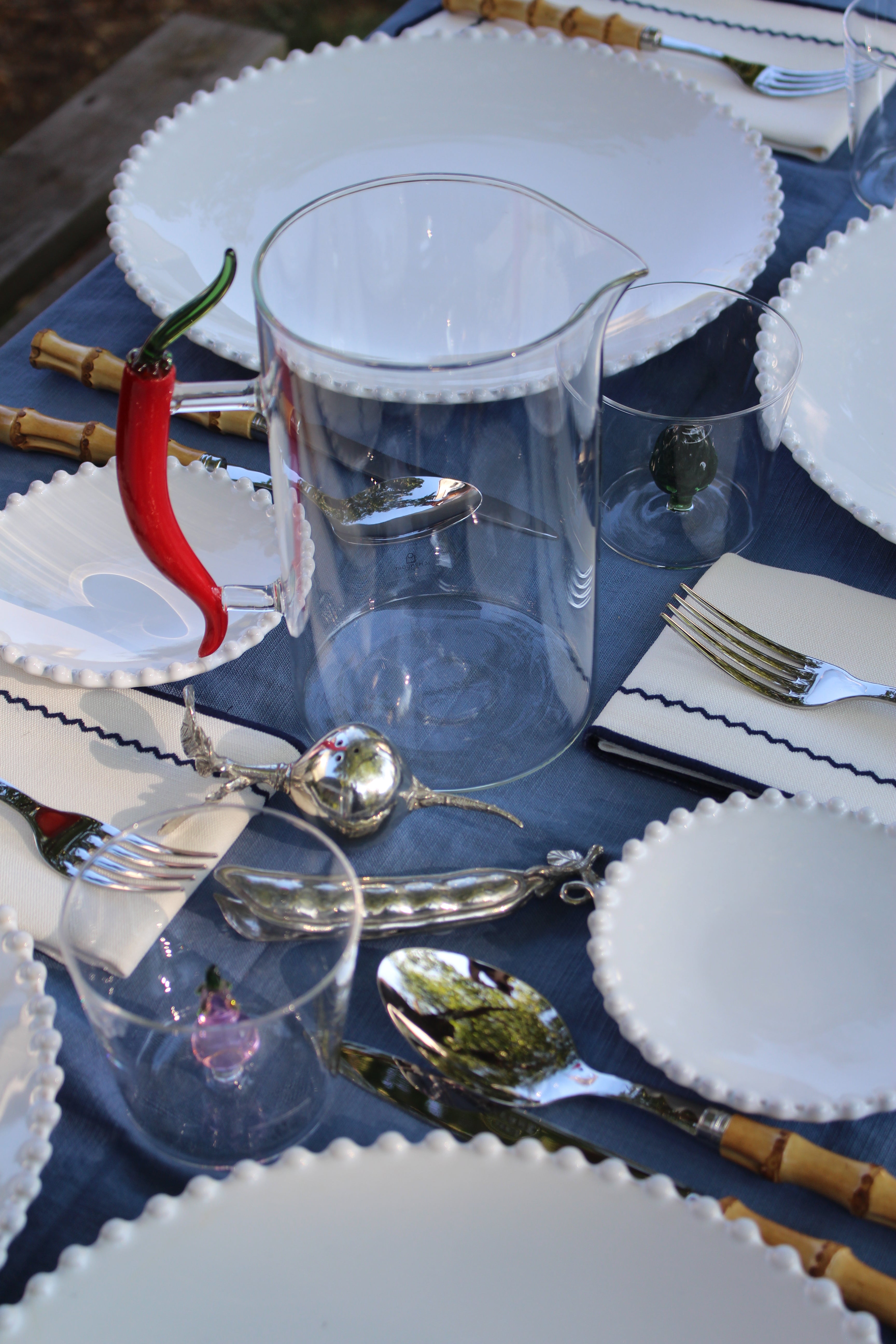 A styled outdoor dining table setting featuring a clear glass pitcher, clear drinking glasses, white scalloped plates, silver cutlery, and a blue tablecloth.