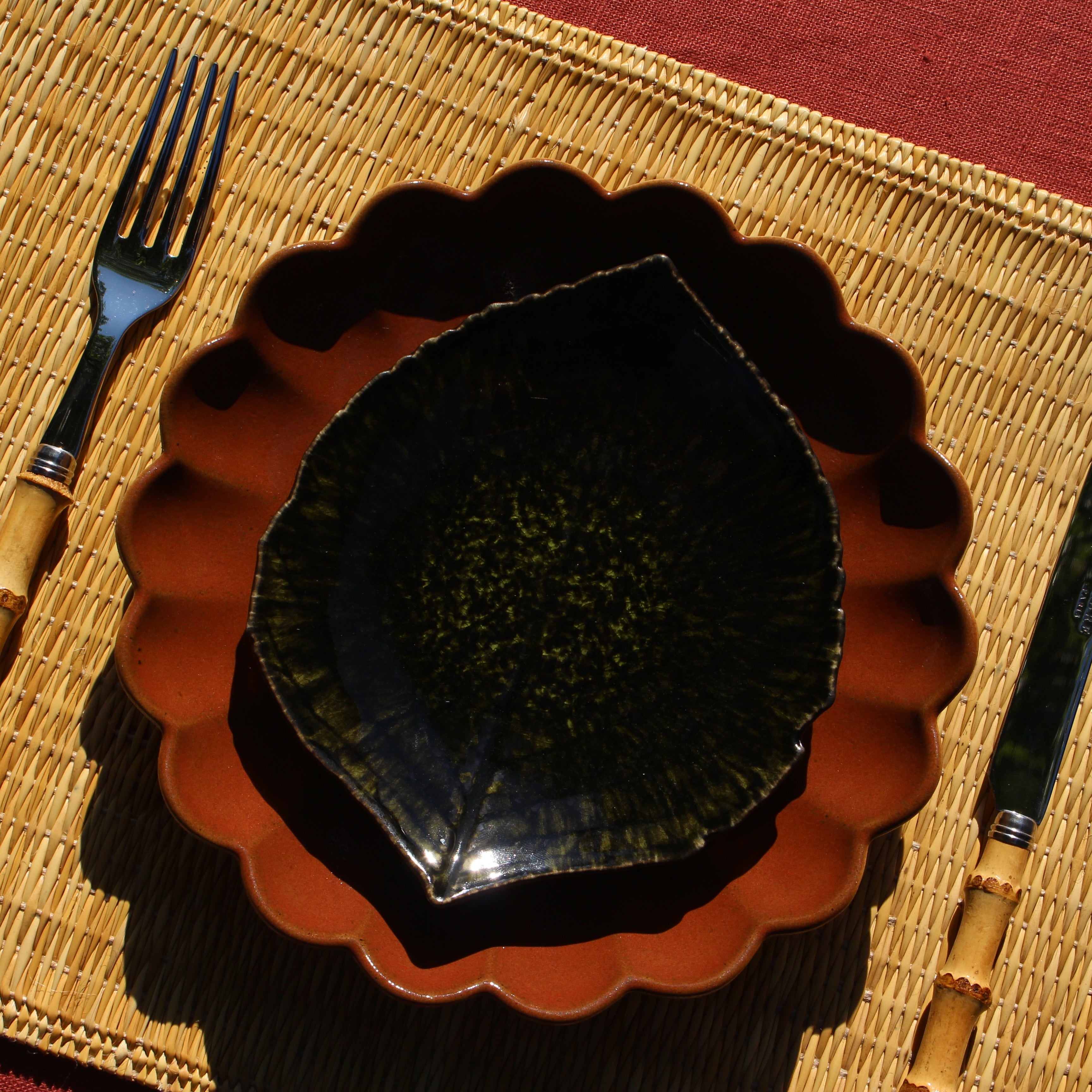 A ceramic leaf-shaped bowl with a dark green glazed interior and a terracotta-colored fluted exterior, styled on a woven placemat with a fork and knife on a red tablecloth.