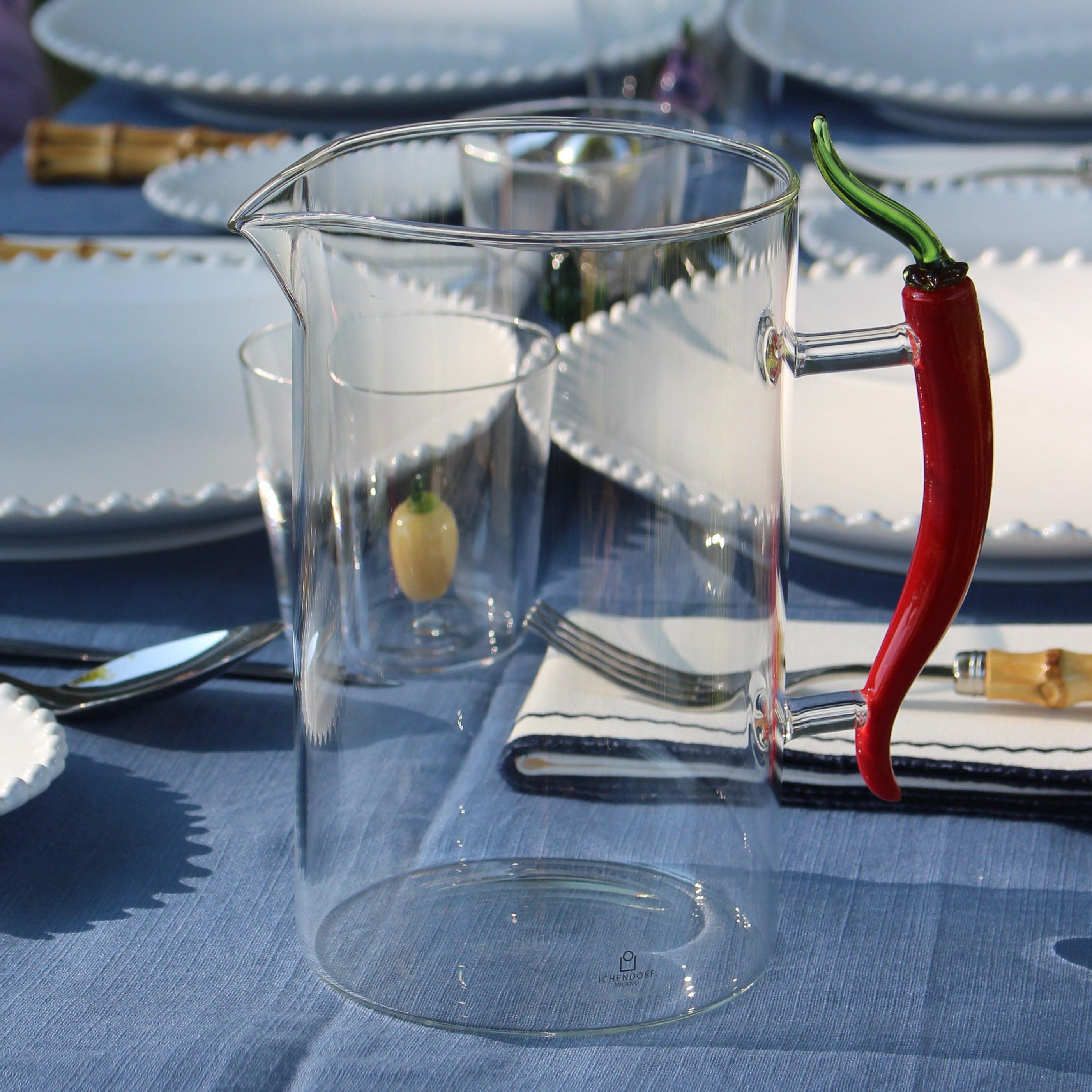 A clear glass pitcher with a handle, decorated with a red chili pepper and a small yellow lemon slice, set on a blue tablecloth next to white plates and gold cutlery.