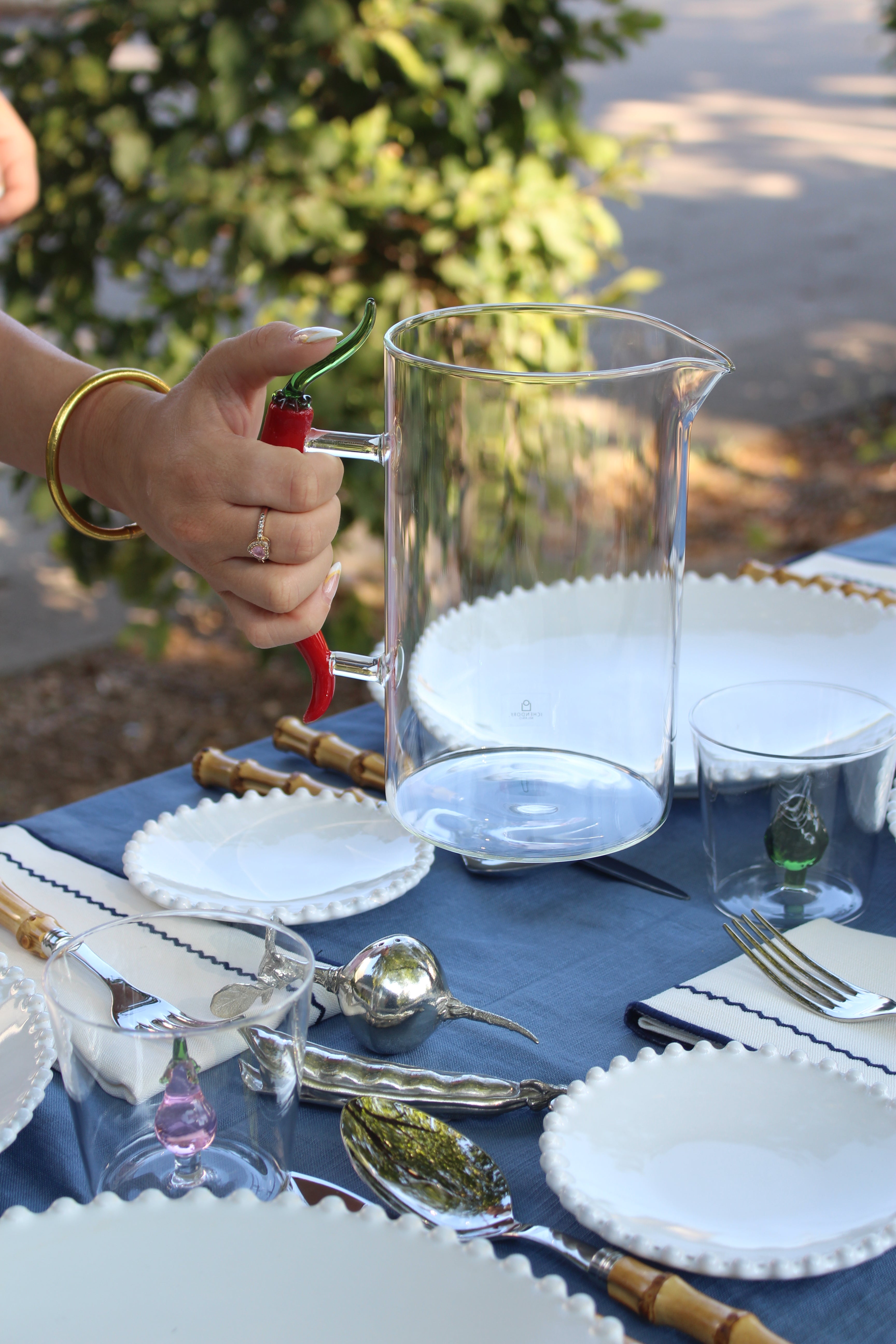 A clear glass pitcher and matching drinking glasses arranged on a blue tablecloth outdoors, with a hand holding a red chili pepper near the pitcher.