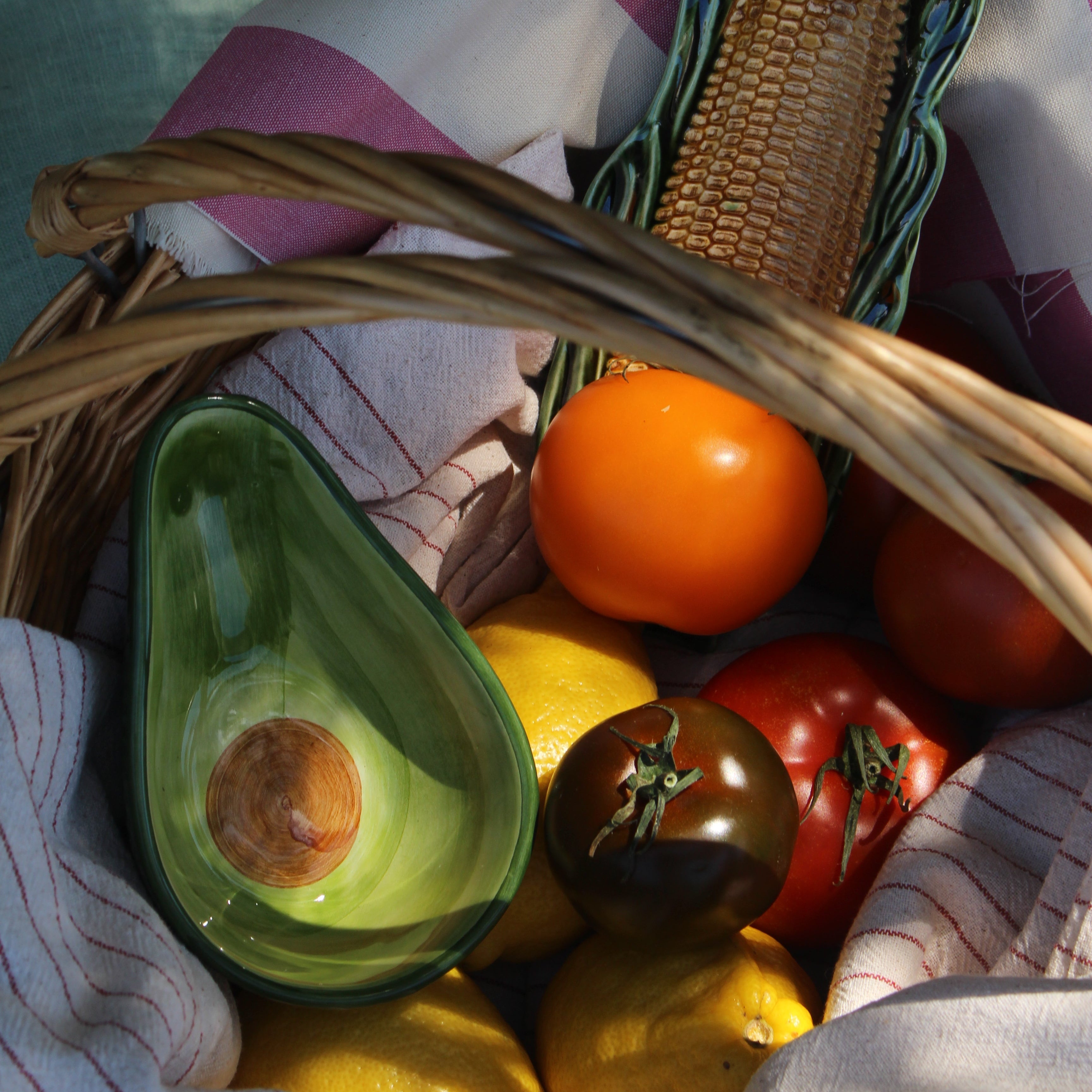 A rustic woven basket filled with fresh produce and decorative ceramic objects, including a green avocado-shaped dish, a corn cob, tomatoes, lemons, and a striped cloth lining.